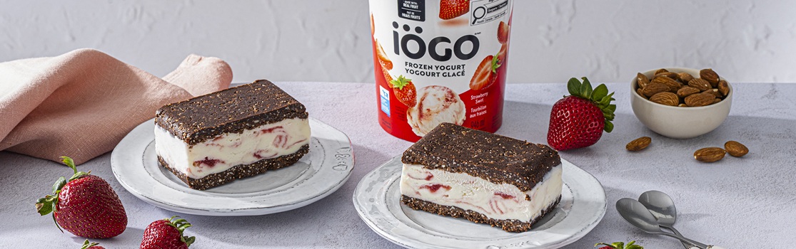 Two plates with No-Bake Brownie Frozen Yogurt Sandwiches made with iÖGO Strawberry Swirl Frozen Yogurt. The dessert is surrounded by fresh strawberries, almonds in a small bowl, and a tub of frozen yogurt in the center.